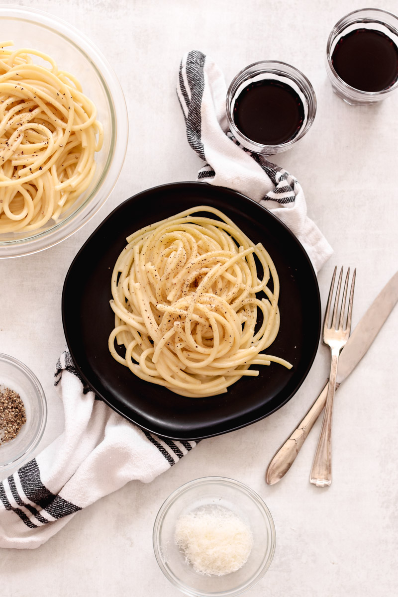overhead image of pasta on a black plate with cups of a dark drink and a fork and knife.
