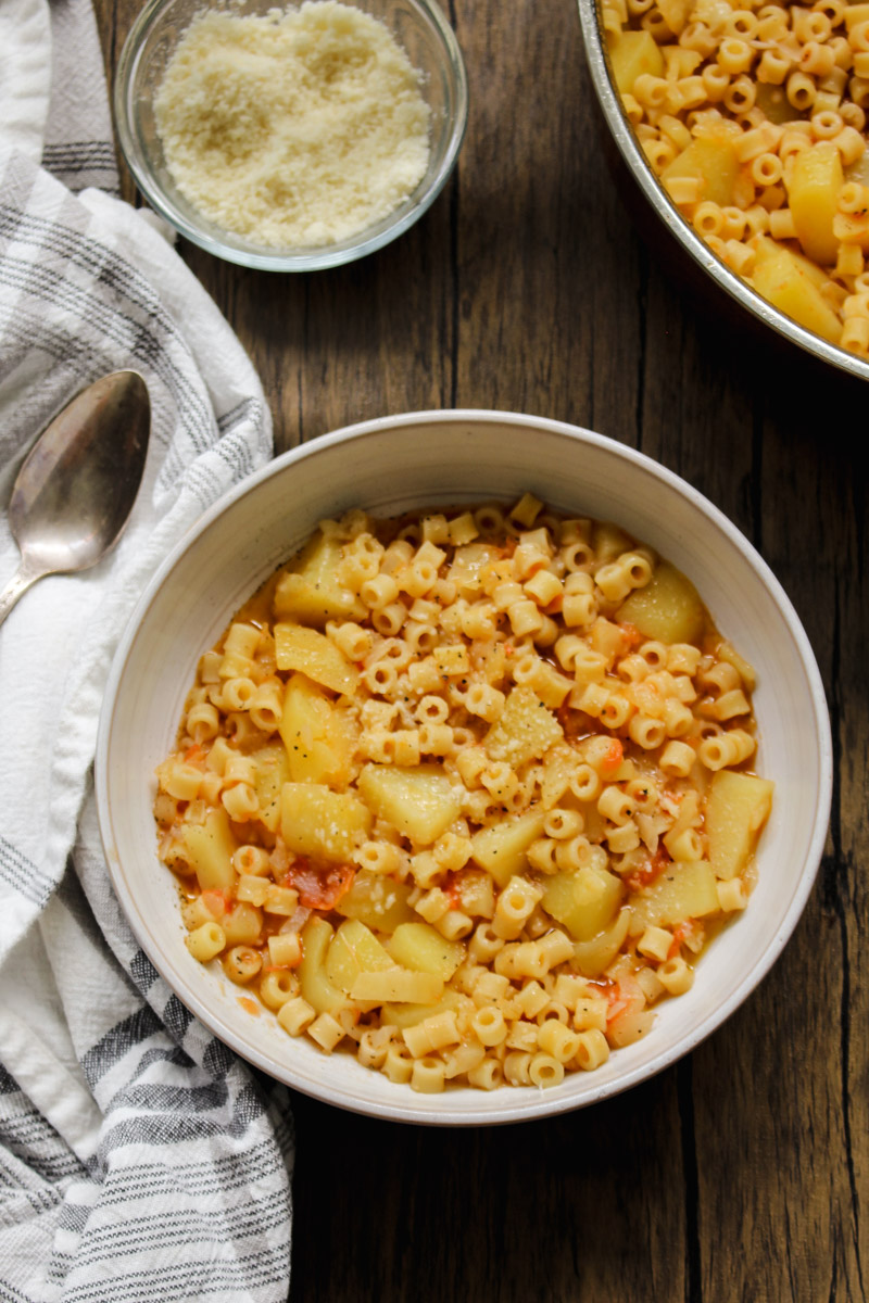 overhead image of pasta with potatoes in a white bowl with a napkin in background with a spoon and grated cheese in a glass bowl. 