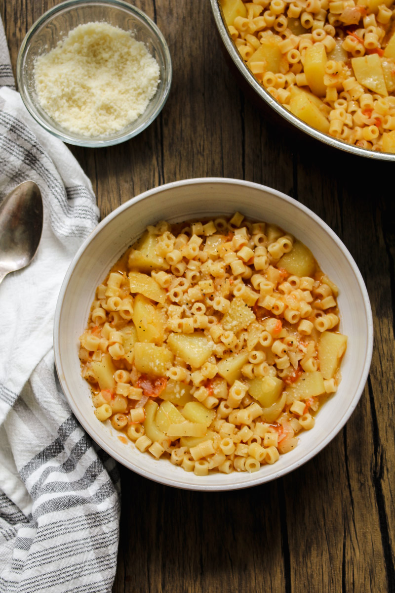 overhead image of pasta and patate in a white bowl on a wood background with a napkin and cheese in a small bowl.