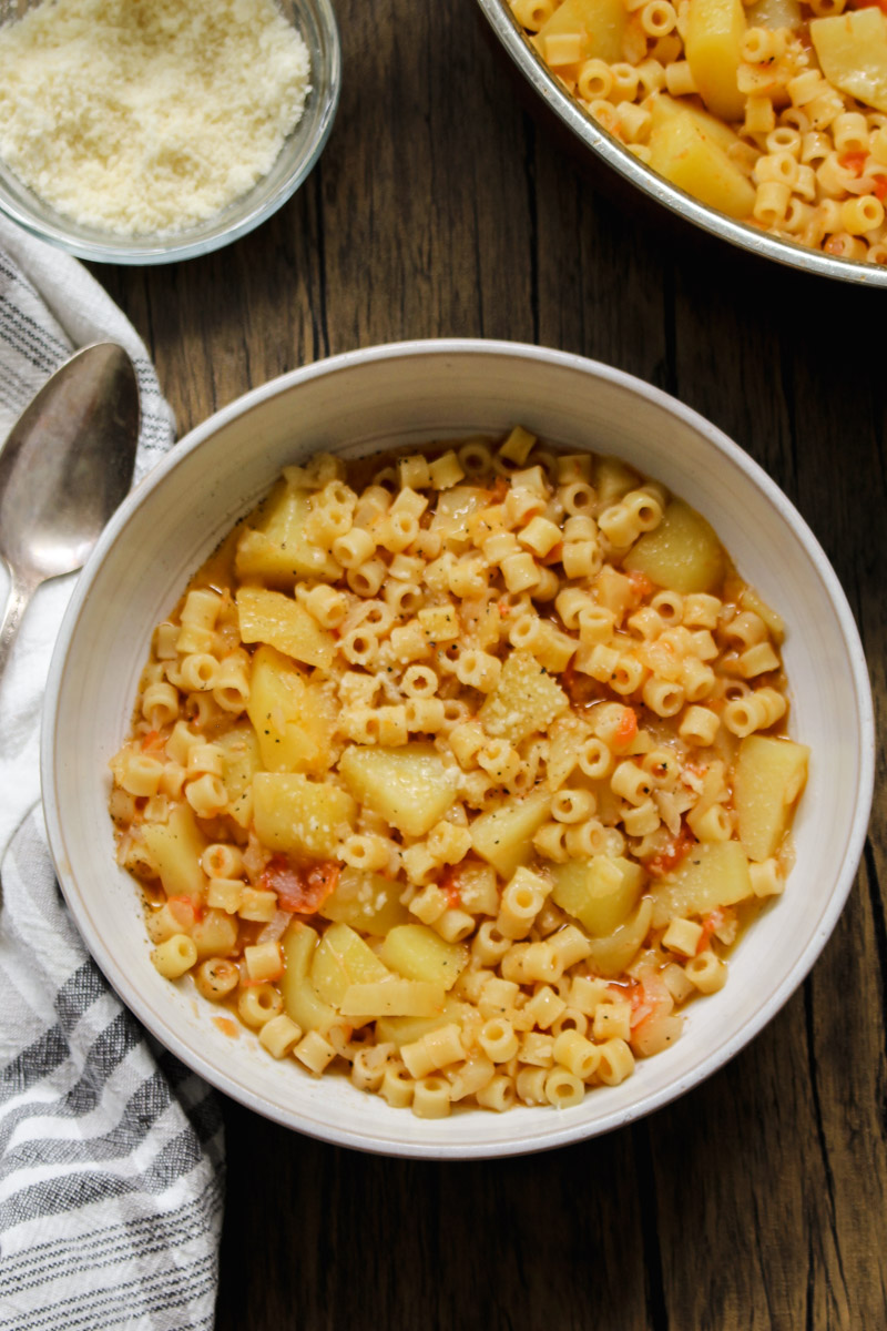 close up image of pasta with potatoes in a white bowl with a napkin in background with a spoon and grated cheese in a glass bowl. 