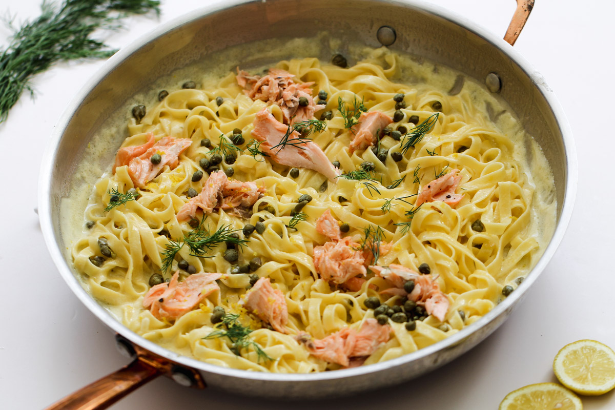 close up image of creamy salmon pasta in a pan with lemon and dill in the background. 