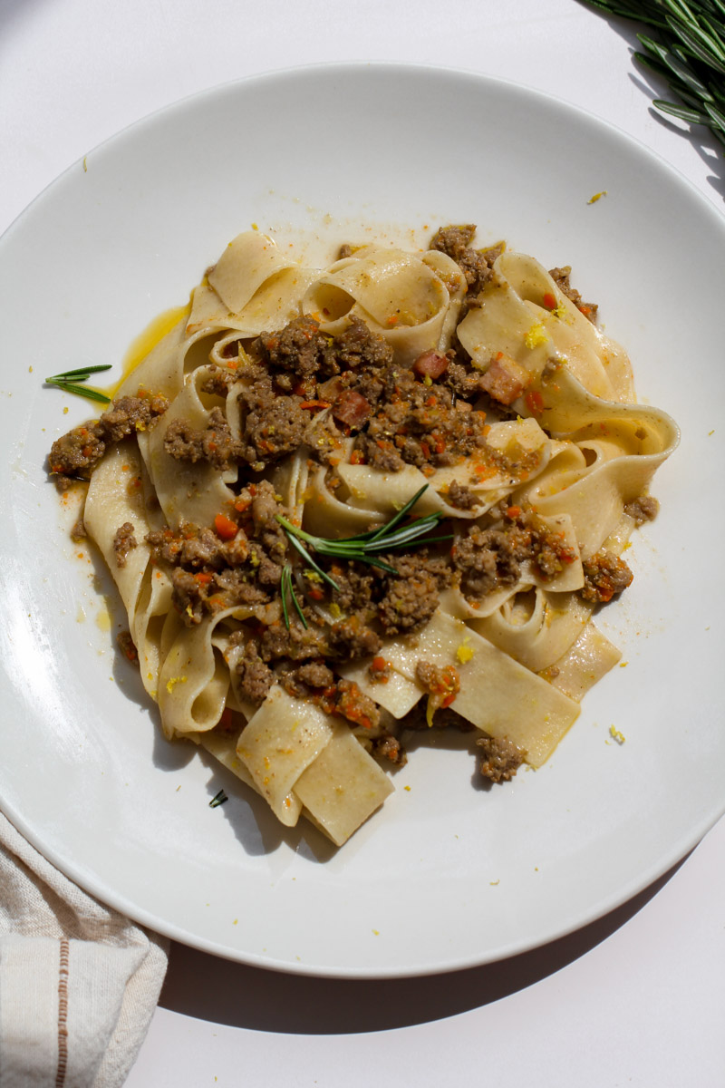 close up image of a pasta with white ragu sauce on a white plate with rosemary in the background. 