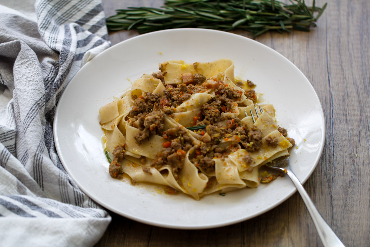 overhead image of a white ragu sauce with pasta on a white plate and fresh rosemary and a towel in the background. 