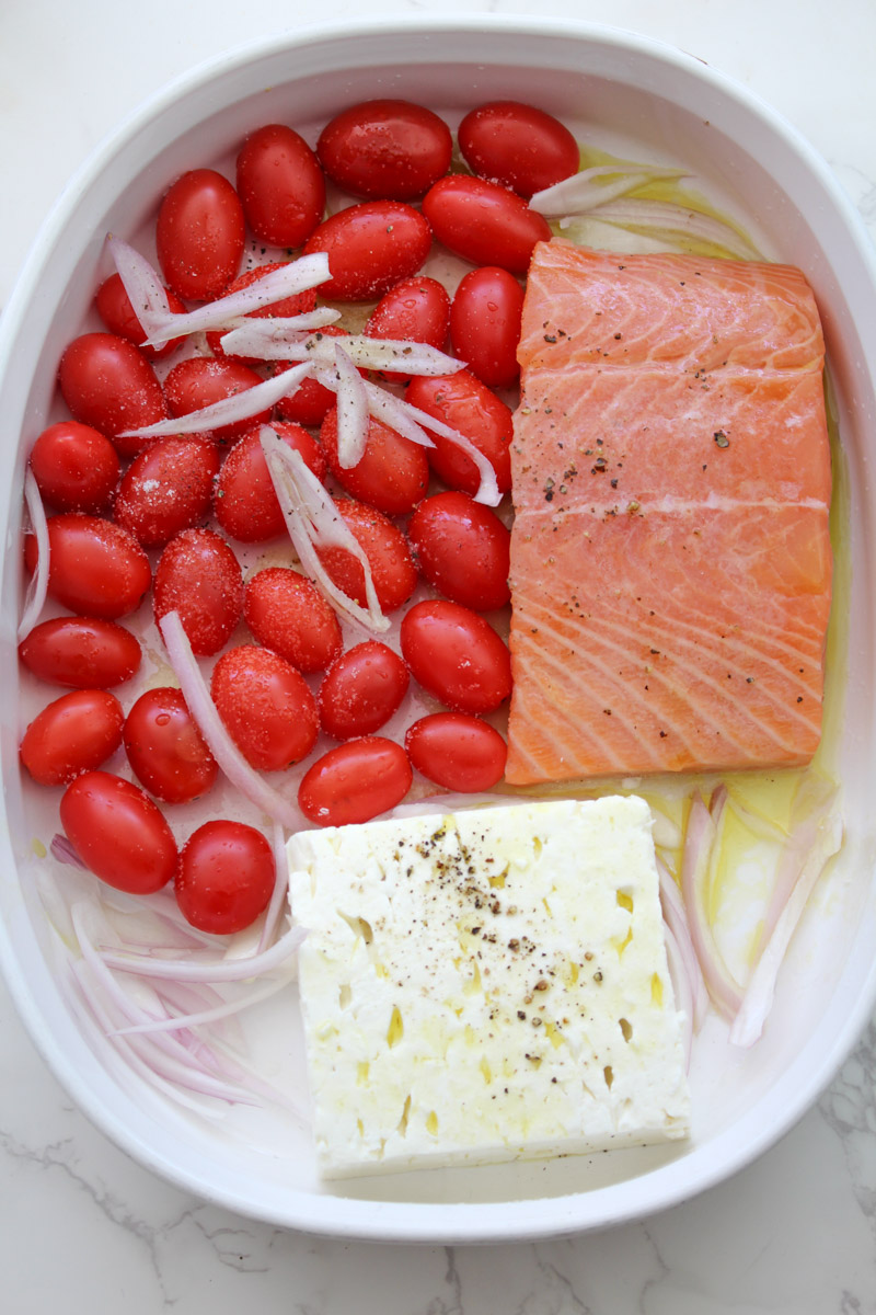 close up image of tomatoes, feta, shallots, salmon in a white casserole. 