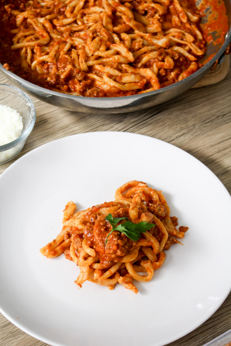 overhead image of pici with bolognese sauce on a white plate and in a skillet on a wood background. 