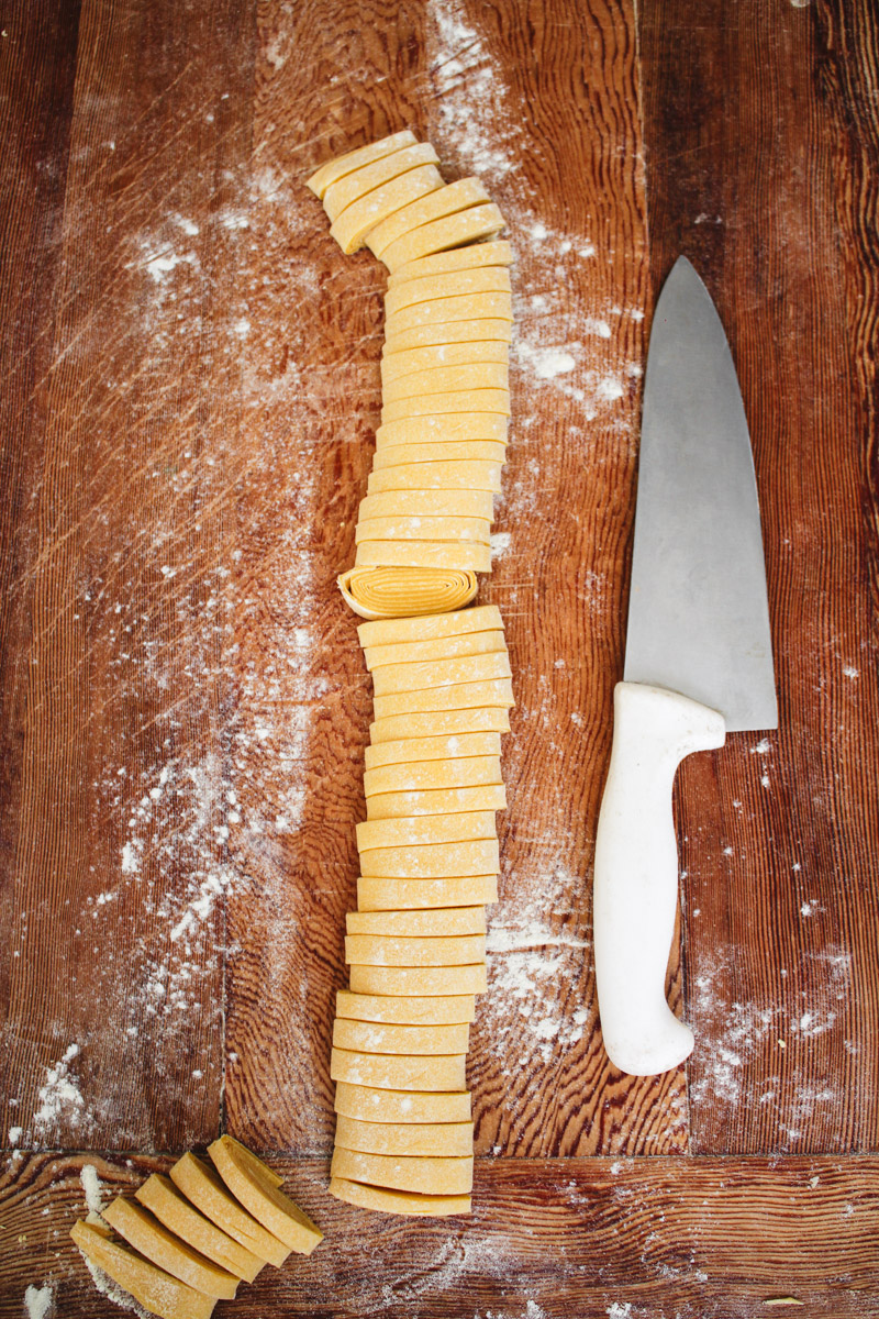 overhead image of slicing dough to make tagliatelle pasta with a knife and a wooden board.
