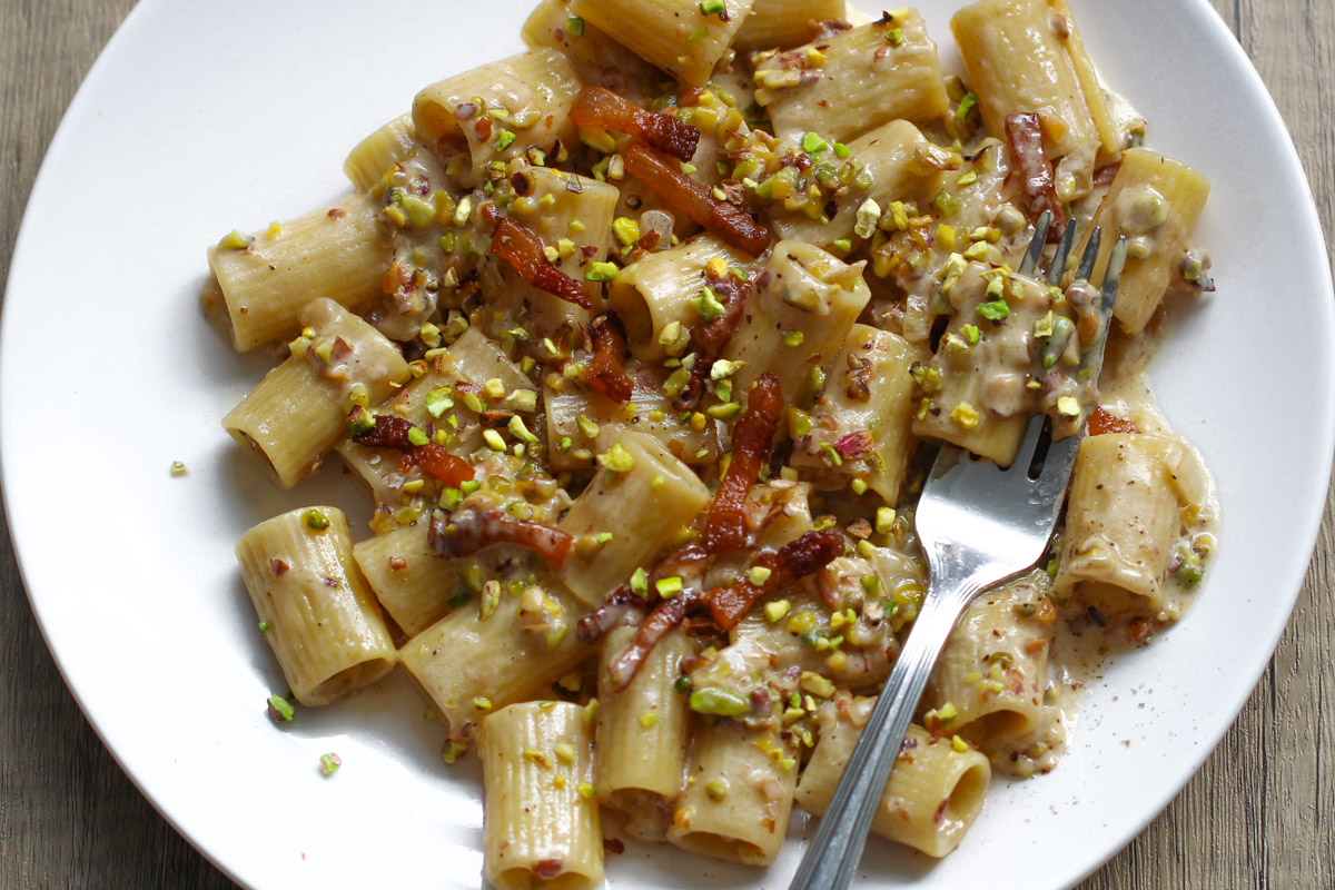 close up image of creamy pistachio pasta on a white plate with a fork and wooden background. 