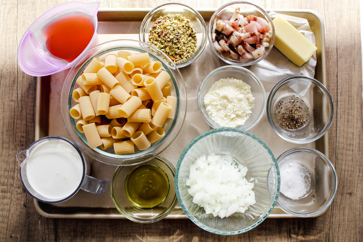 overhead image of ingredients to make creamy pistacho pasta in glass bowls on a baking tray and wooden background. 