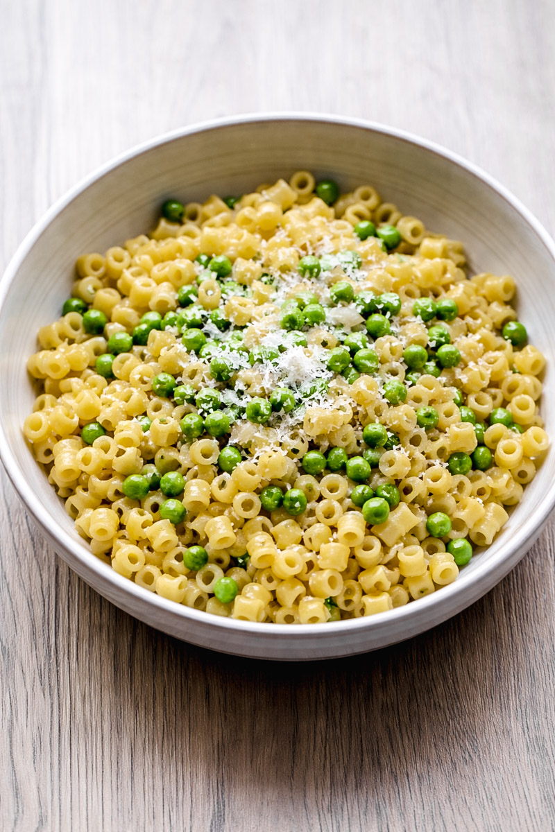 close up image of pasta and peas in a white bowl on a wooden background.
