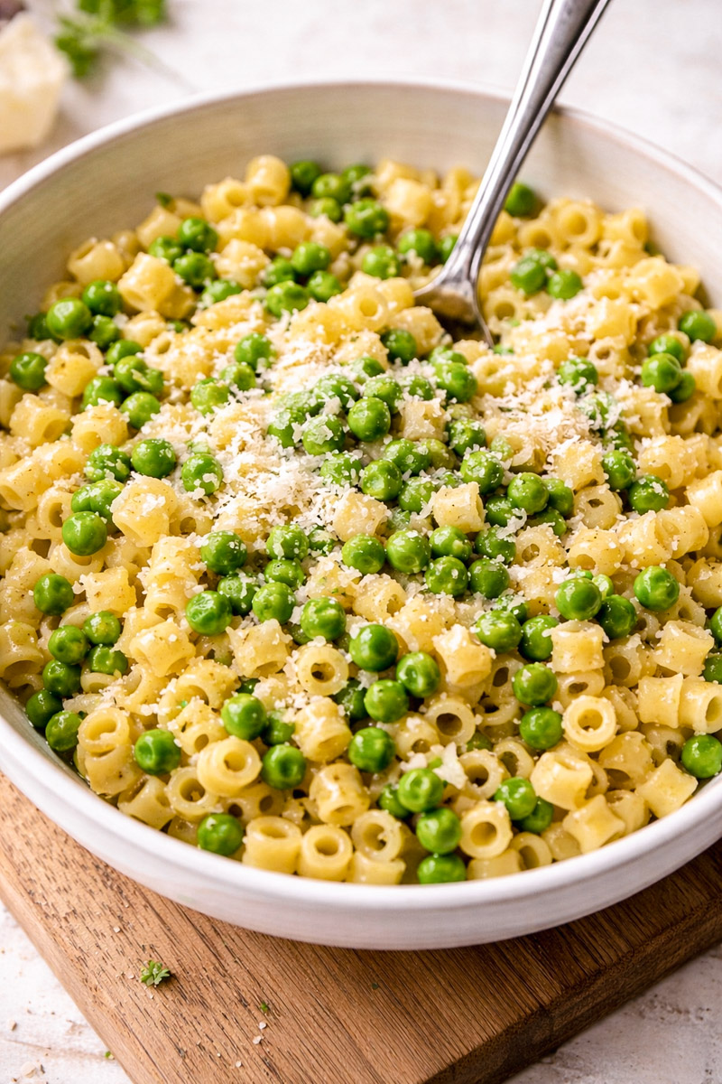 close up image of pasta and peas served in a white bowl on a wooden board, with a fork resting inside.